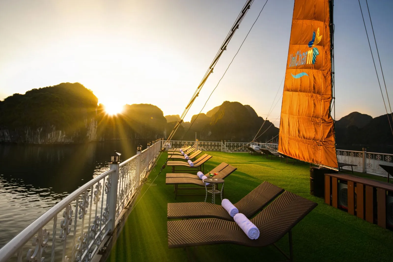 Sun-deck with rows of loungers and rolled towels on artificial grass beside a white railing, an orange sail printed with a logo, and limestone karst islands with the sun setting over calm water.