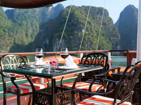 A set dining table with two wine glasses, napkins and a flower, flanked by ornate metal chairs with orange-striped cushions on a wooden boat deck, with green karst limestone peaks across the water.