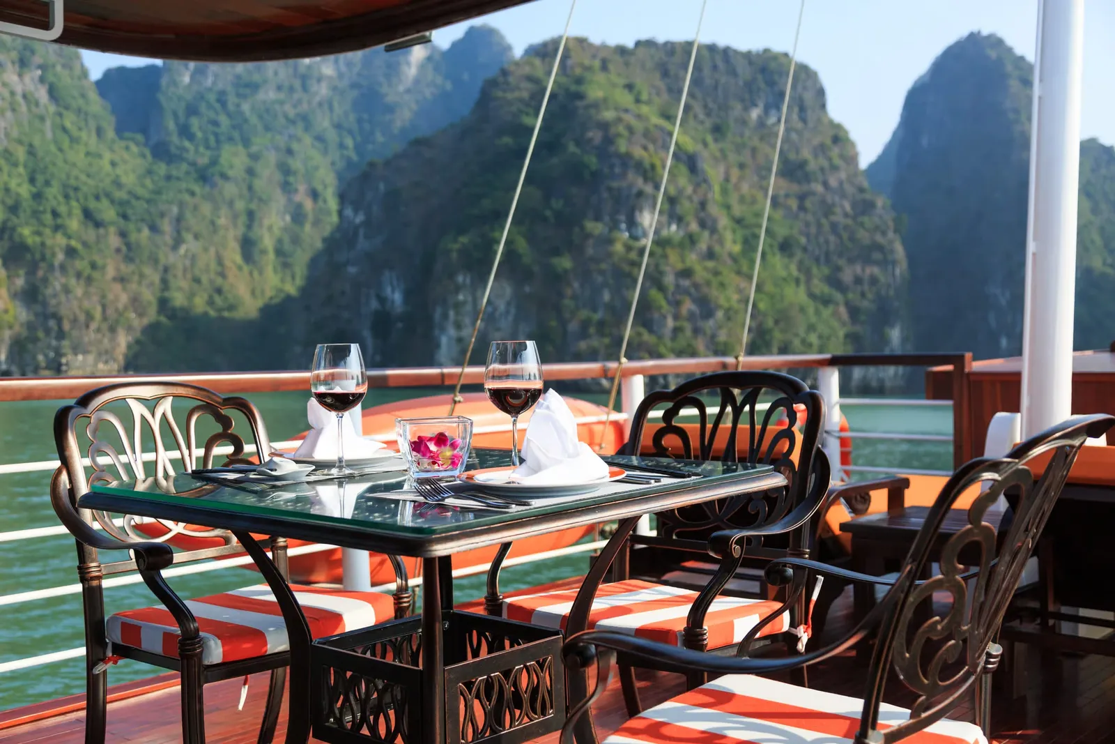 A set dining table with two wine glasses, napkins and a flower, flanked by ornate metal chairs with orange-striped cushions on a wooden boat deck, with green karst limestone peaks across the water.