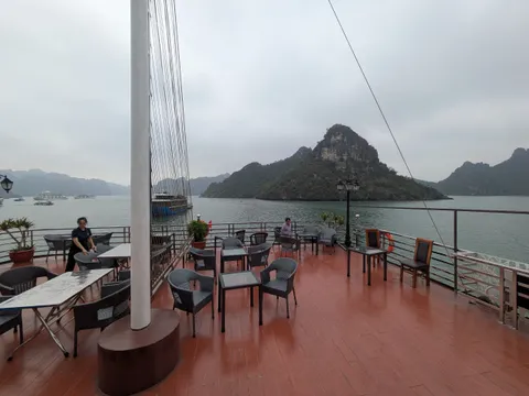 A boat deck with orange flooring, tables and chairs, a white mast, and limestone karst islands in the background across calm water.
