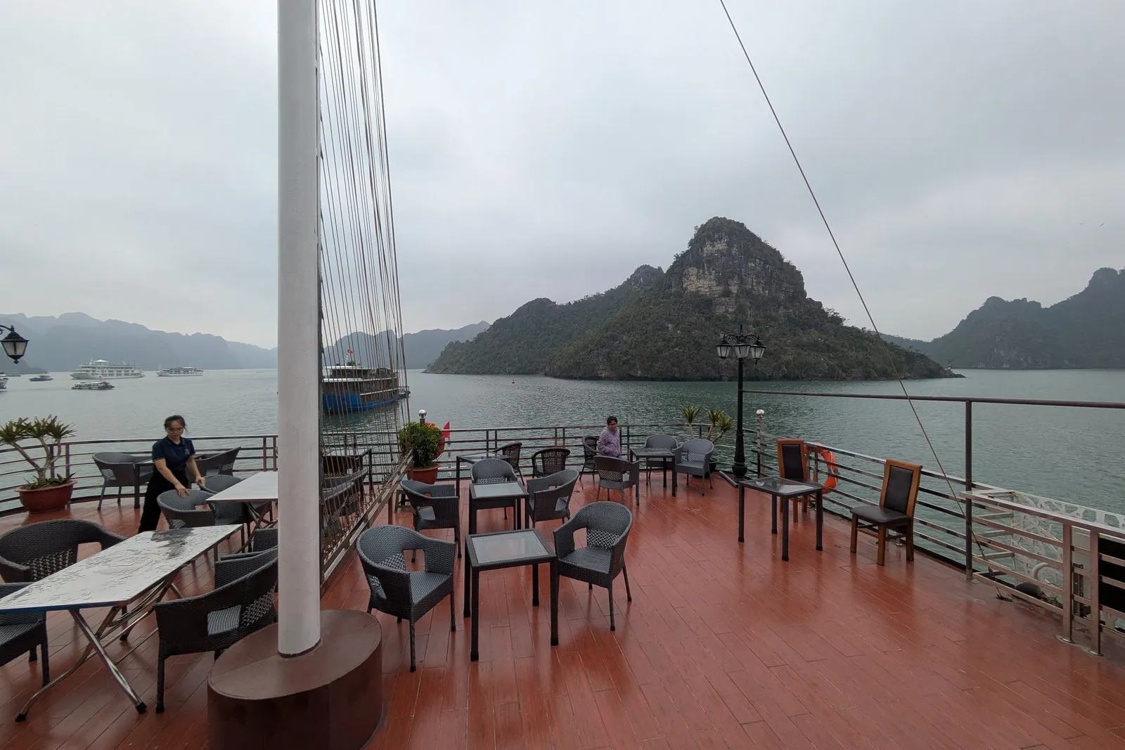 A boat deck with orange flooring, tables and chairs, a white mast, and limestone karst islands in the background across calm water.