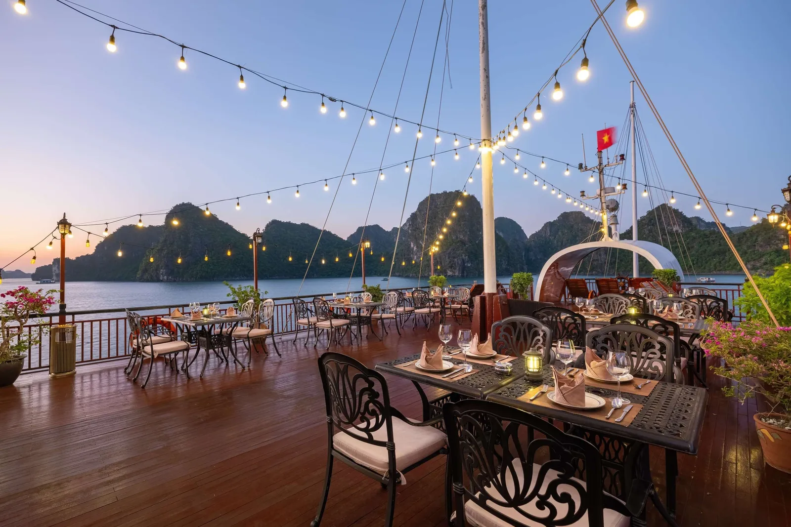 An outdoor dining deck on a boat with set tables, string lights, and limestone karst mountains in the background at dusk.