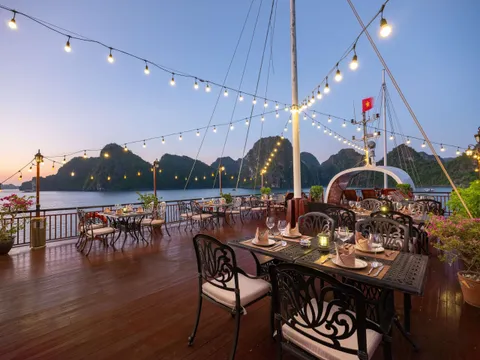 An outdoor dining deck on a boat with set tables, string lights, and limestone karst mountains in the background at dusk.