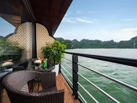 A private balcony on a boat with wicker chairs, a glass table with two cocktails and flowers, a potted plant and a railing, overlooking green water and forested limestone karst islands.