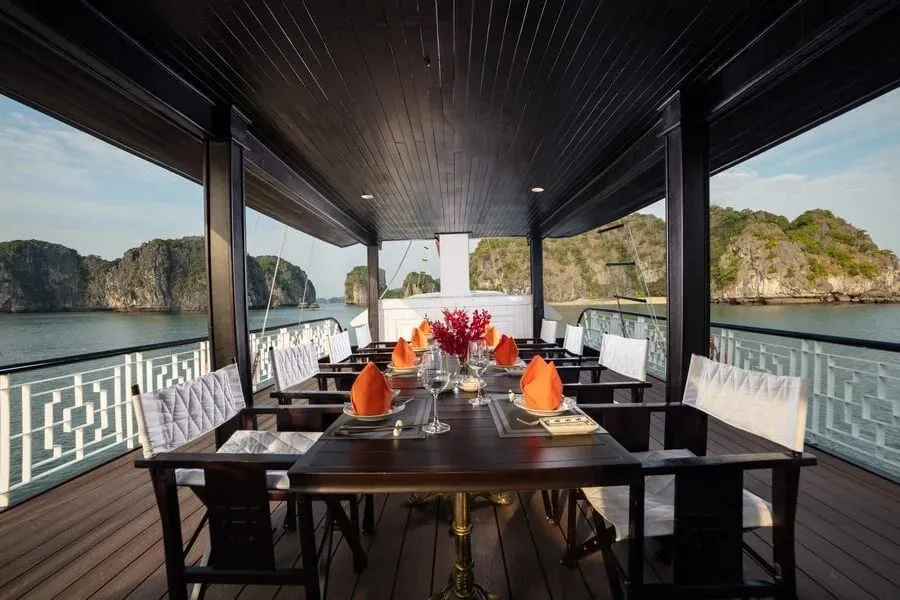 A covered upper deck with a dark wooden dining table set with orange napkins, glasses and red flowers, white railings on both sides, and limestone karst islands across calm water in the background.