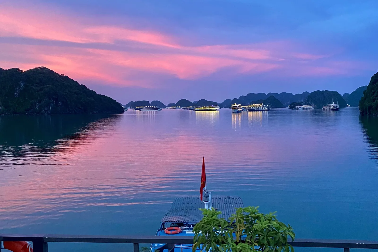 A boat deck with railings, lounge chairs, a potted plant, and Vietnamese flag at sunset with limestone karsts and illuminated cruise ships in the bay.