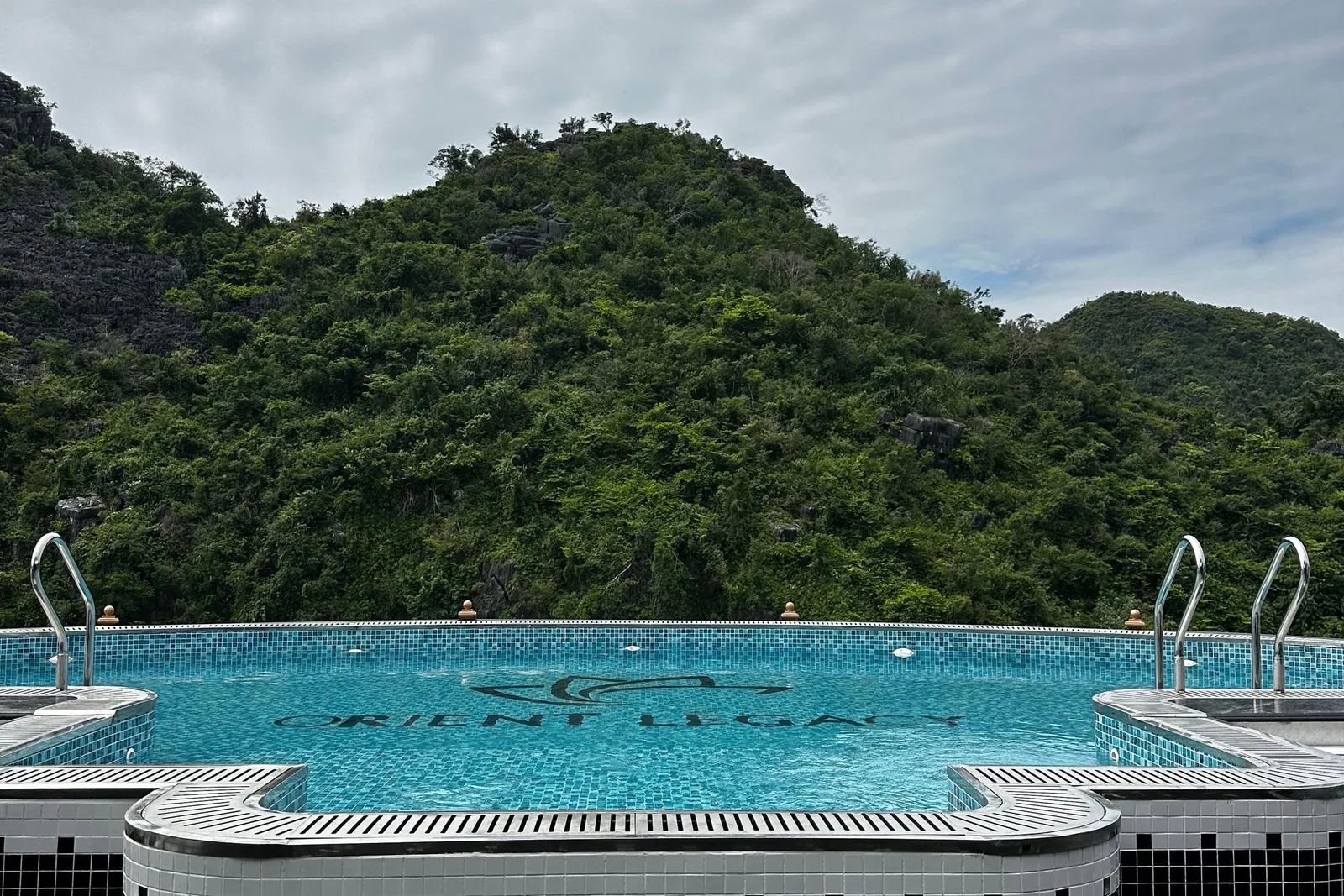 A tiled infinity pool on a boat's upper deck with 'Orient Legacy' and a logo set into the pool floor, facing a forested limestone hill under a cloudy sky.