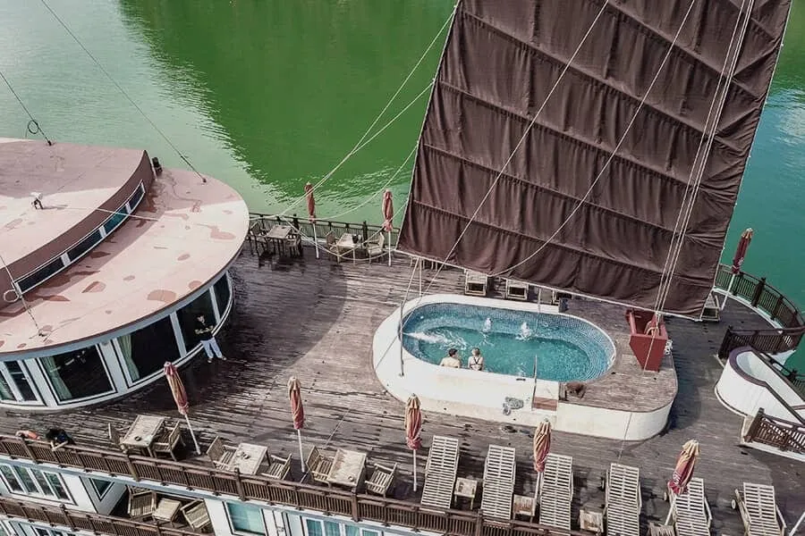 High-angle view of a wooden cruise ship sundeck with a small jacuzzi holding two people, loungers, closed umbrellas, tables, and a large brown junk sail above green water.