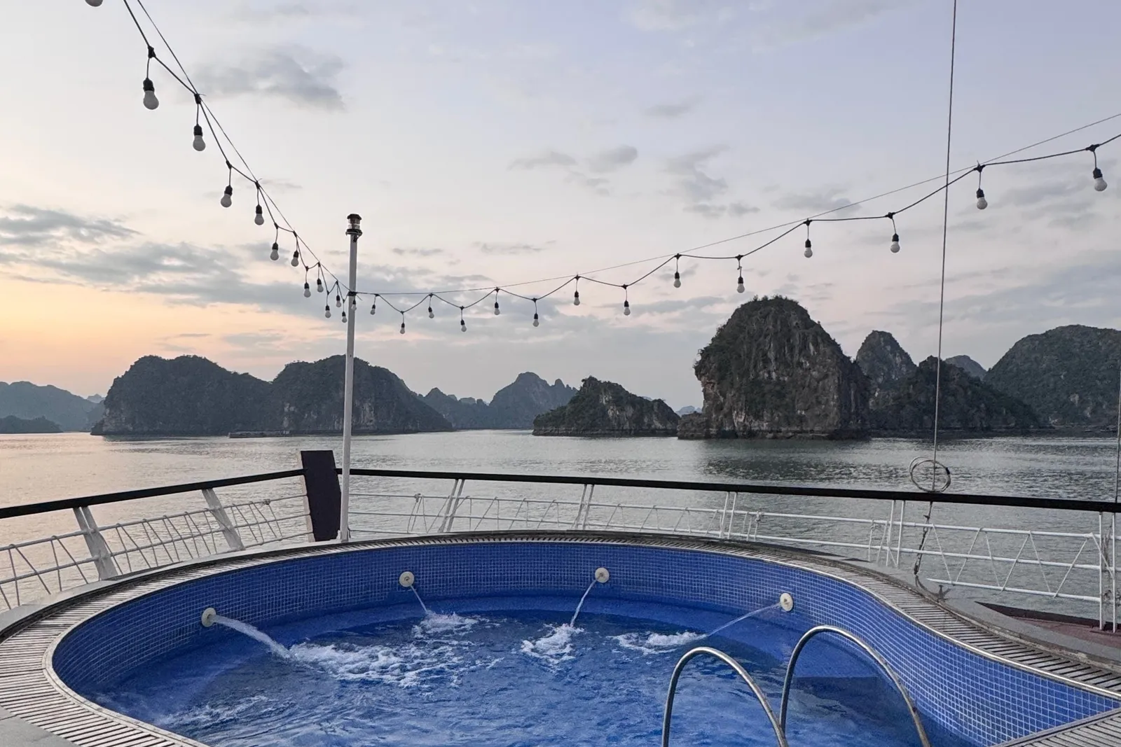 A blue tiled jacuzzi pool on a wooden boat deck at sunset with limestone karst islands in the background.