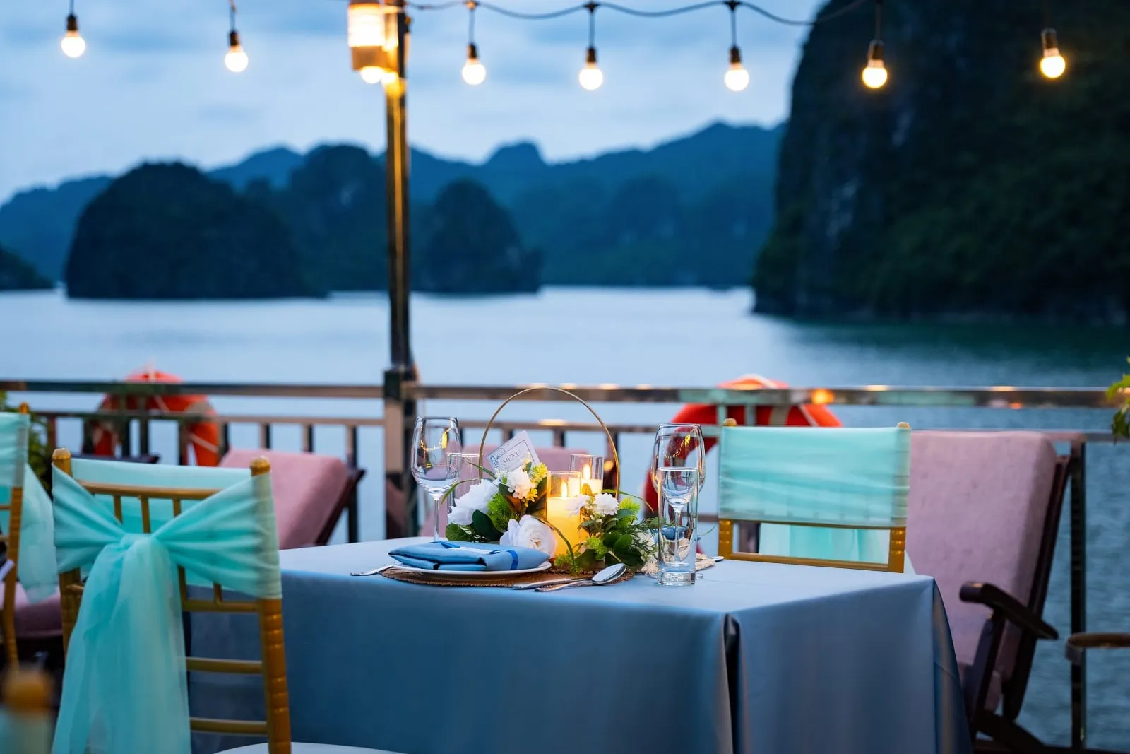 A dusk dinner table set with a blue cloth, flowers, candles and glasses on a boat deck with teal-sashed chairs, string lights, life rings on the railing, and limestone karsts rising from the bay behind.