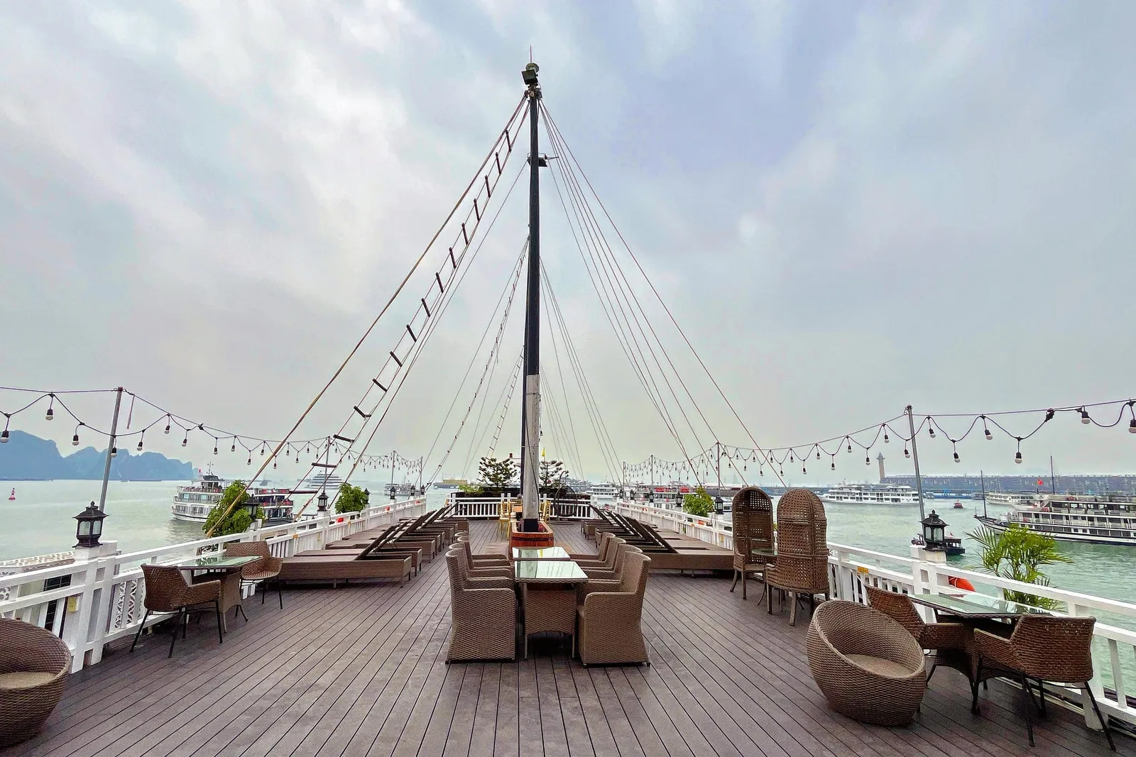 A wooden deck on a cruise boat with wicker furniture, a tall mast with rigging, string lights, and limestone karsts visible in the bay behind.