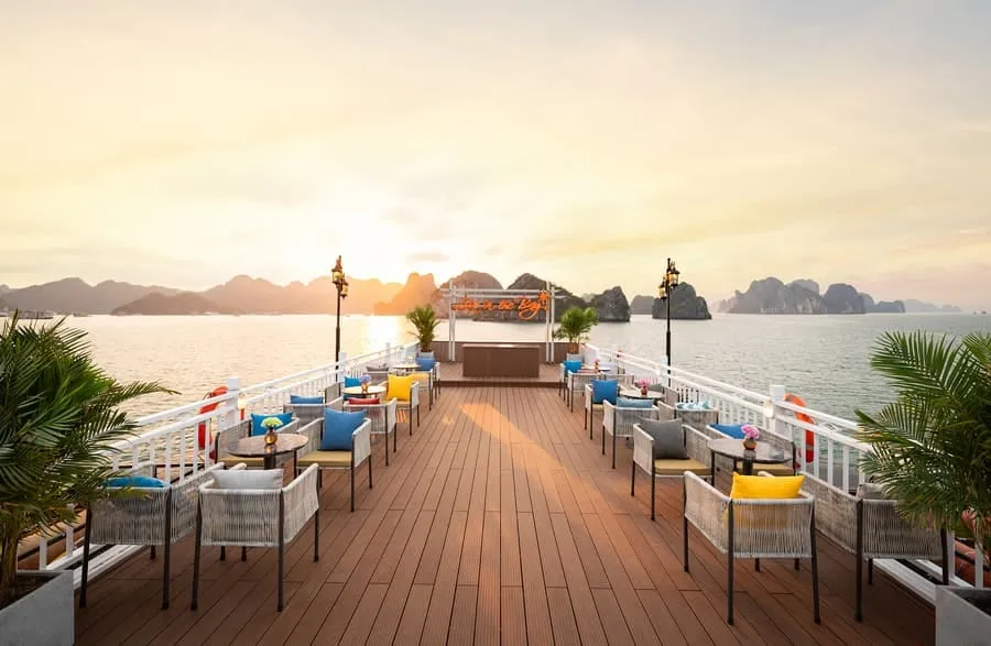A long timber sundeck lined with wicker tables and chairs facing a bay at sunset.
