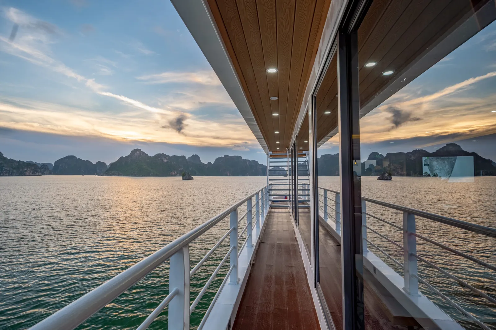 A wood-floored outdoor walkway along the side of the boat with a white railing, looking out over the bay at sunset.