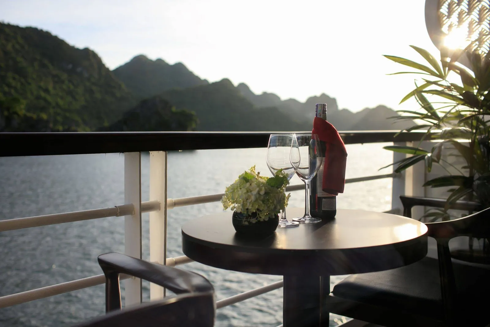 A small round table on a boat deck with two wine glasses, a wine bottle with a red napkin and a pot of white flowers, beside a white railing overlooking calm water and forested limestone hills in warm backlight.