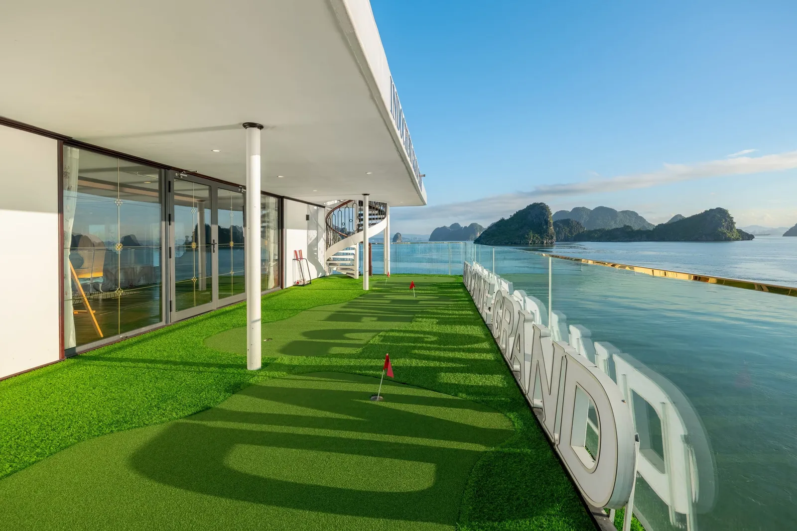 A cruise ship deck with artificial grass putting green, white railings with large letters, glass cabin walls, and limestone karst islands in calm water.