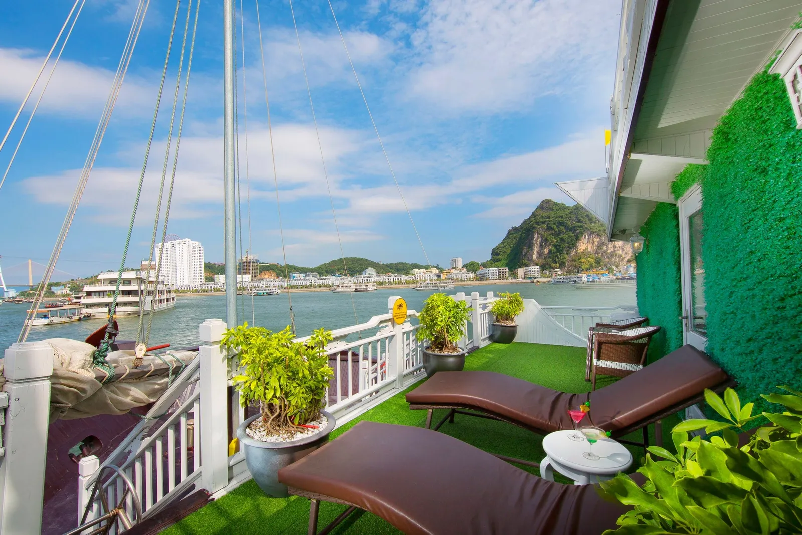 Sun deck of a wooden junk with artificial grass, two brown loungers, potted plants, cocktails on a small table and rigging, overlooking the bay, a limestone hill, a bridge and the waterfront city.
