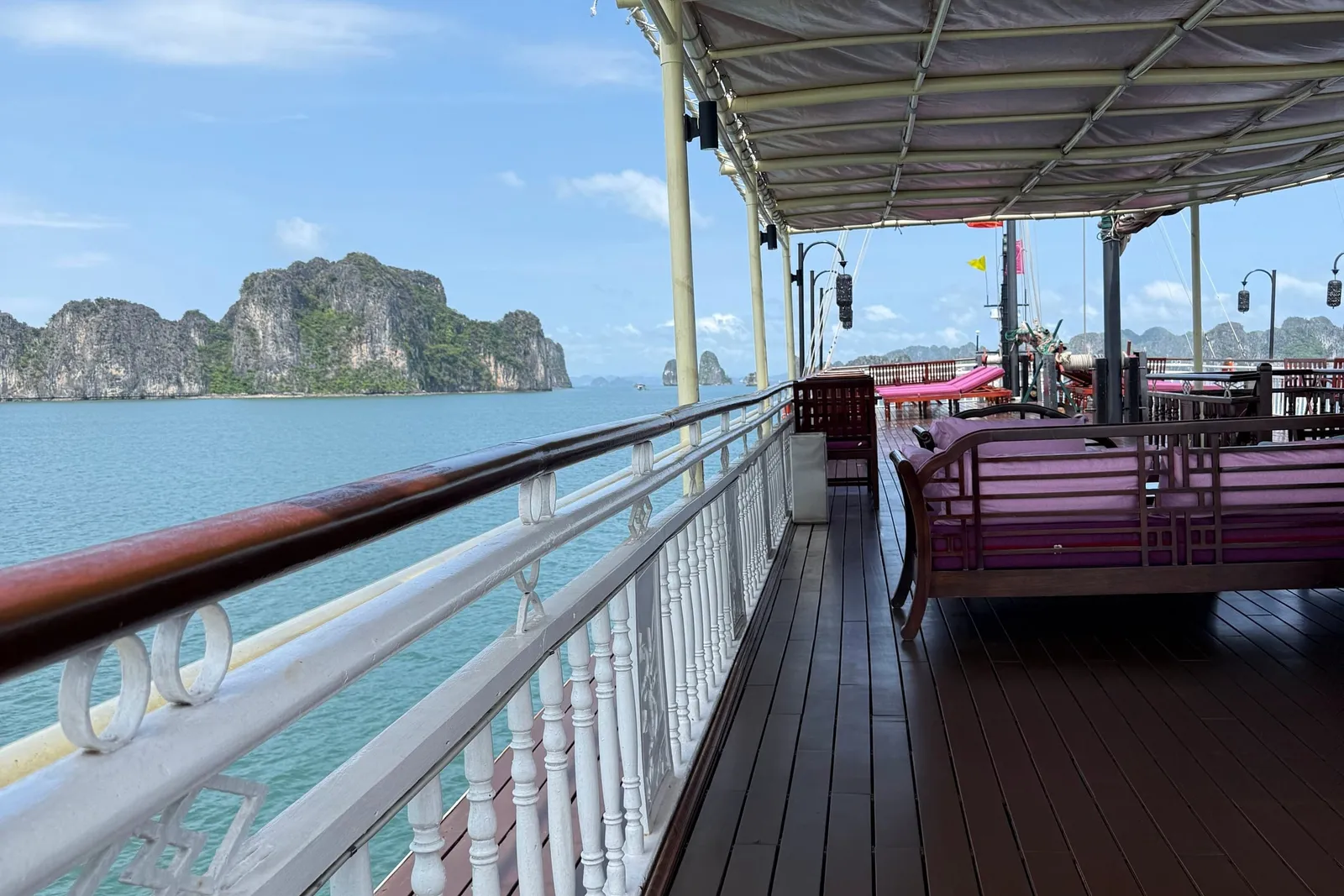 A covered wooden upper deck with white balustrade, pink-cushioned wooden daybeds and loungers, overlooking turquoise water and limestone karst islands under a blue sky.