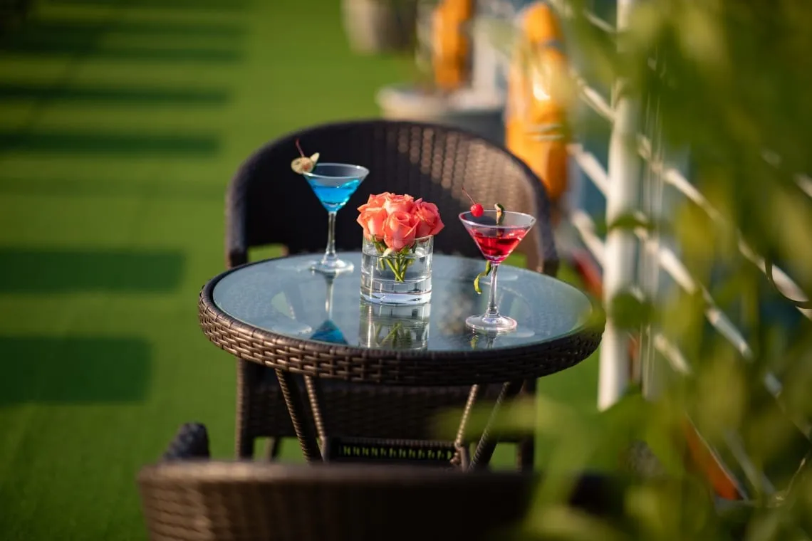A round wicker table with a glass top holds a blue cocktail, a red cocktail and a small vase of pink roses, with wicker chairs on green artificial turf and a white railing with orange life jackets blurred in the background.