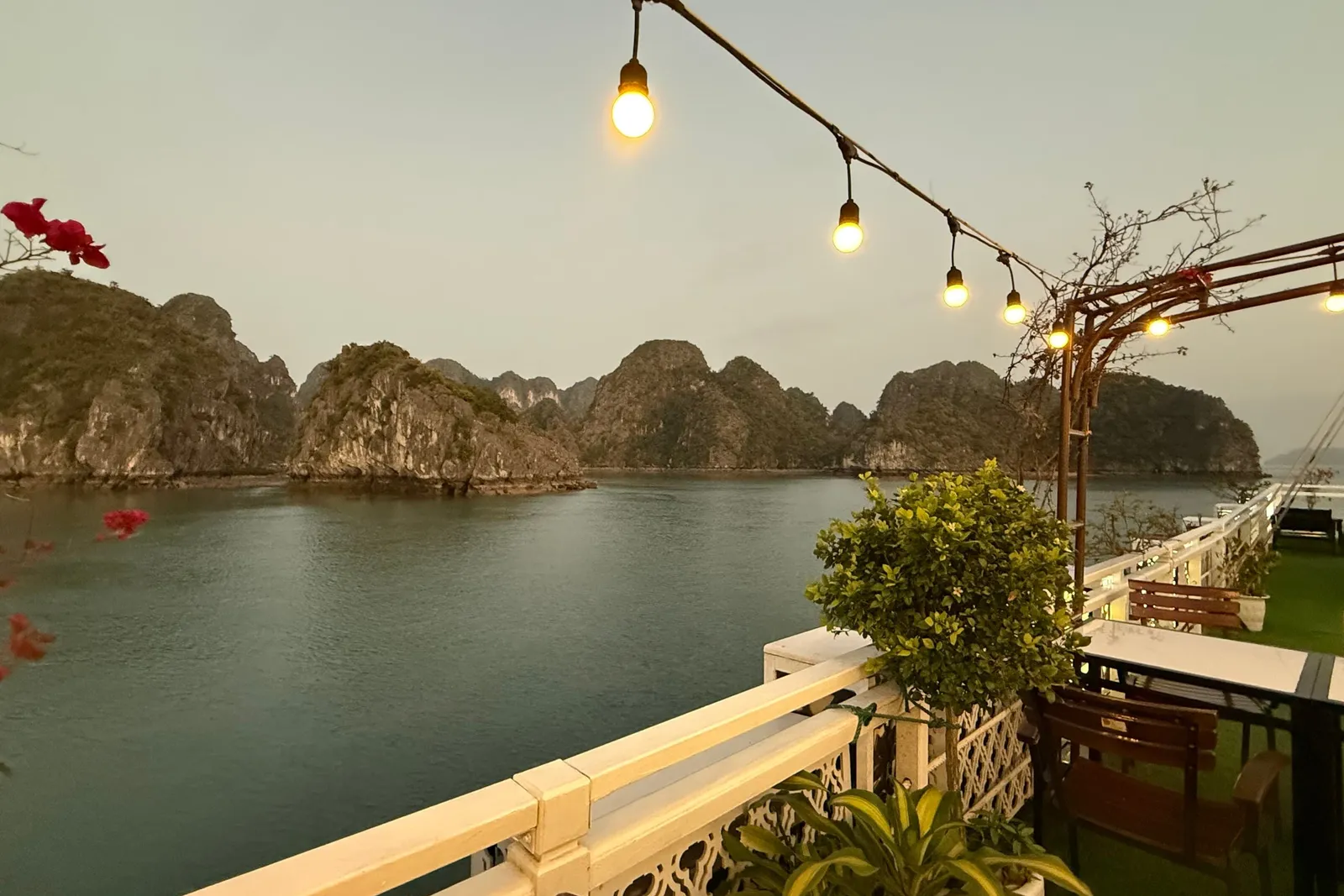 A boat deck with string lights, wooden railings, potted plants, and limestone karst islands in the water at dusk.