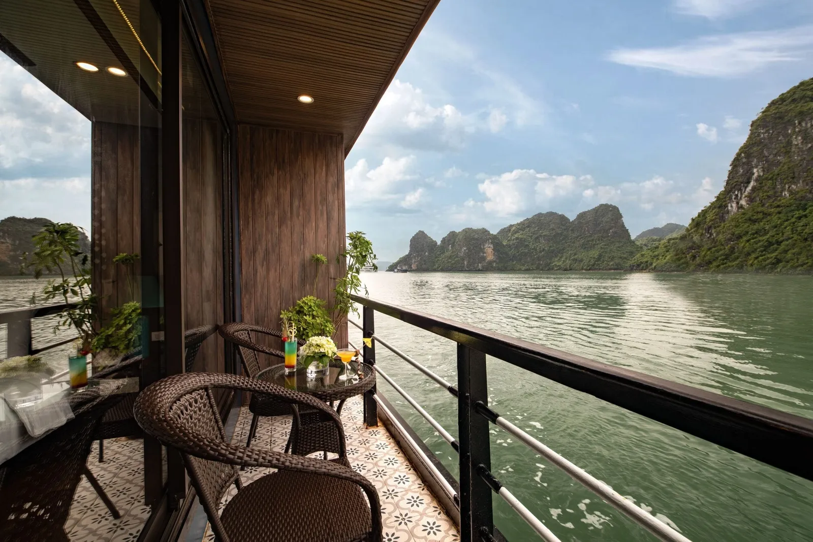 A private cabin balcony with wicker chairs, a small table with drinks and flowers, and a railing overlooking green water and karst limestone islands.