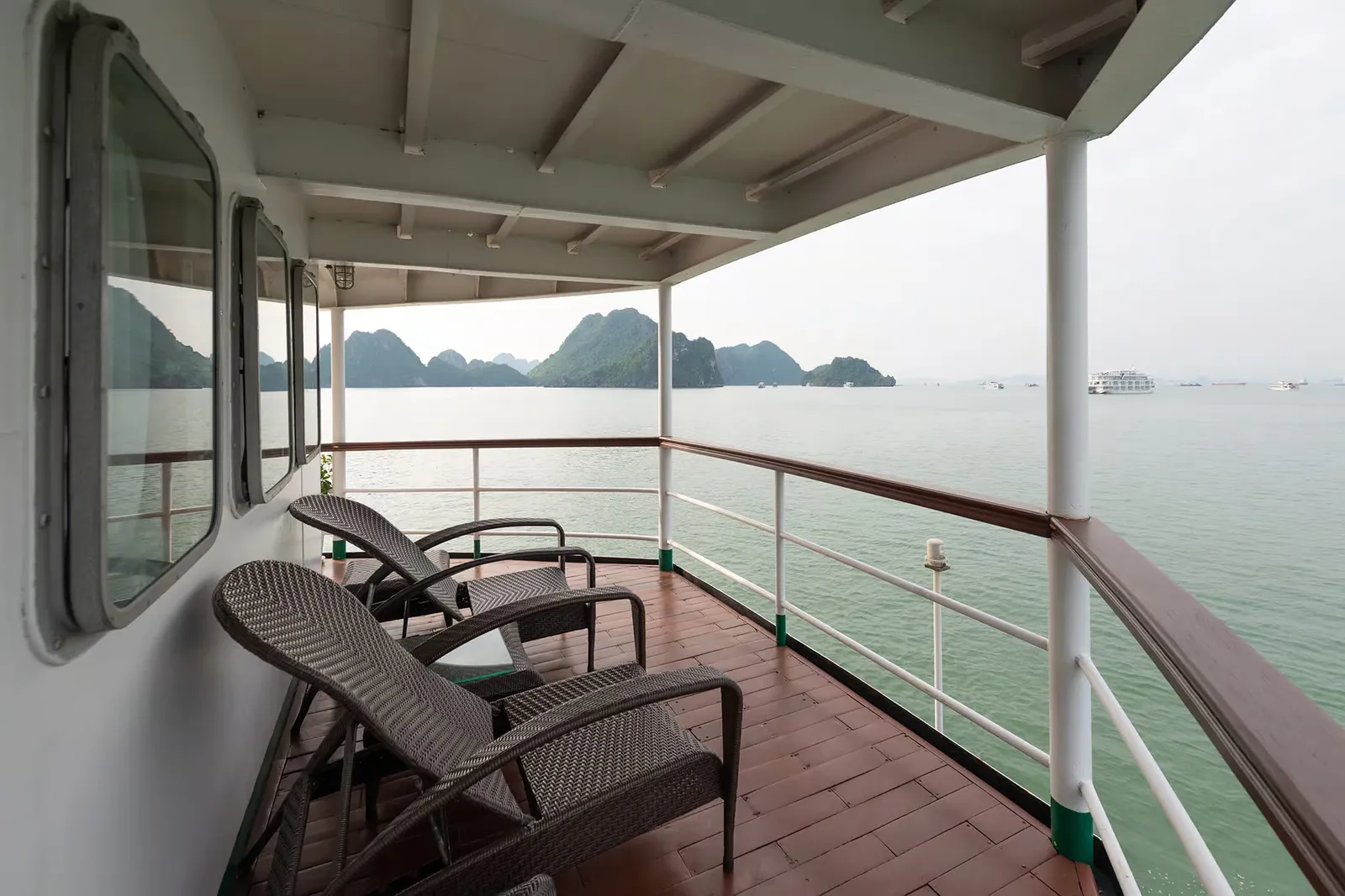 A covered wooden side deck with wicker lounge chairs, white railings and portholes, overlooking calm green water, limestone karst islands and a distant white cruise boat.