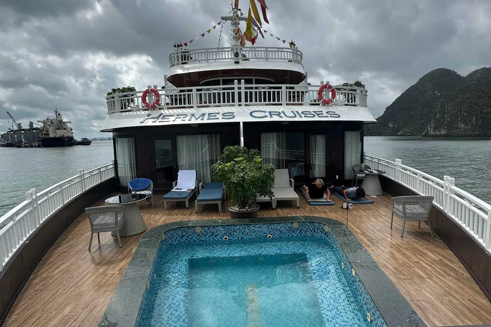 Sun deck of a cruise ship with a small tiled plunge pool, loungers and two people lying down, with 'Hermes Cruises' lettered on the upper deck fascia, under a cloudy sky with a karst island and a cargo ship in the distance.