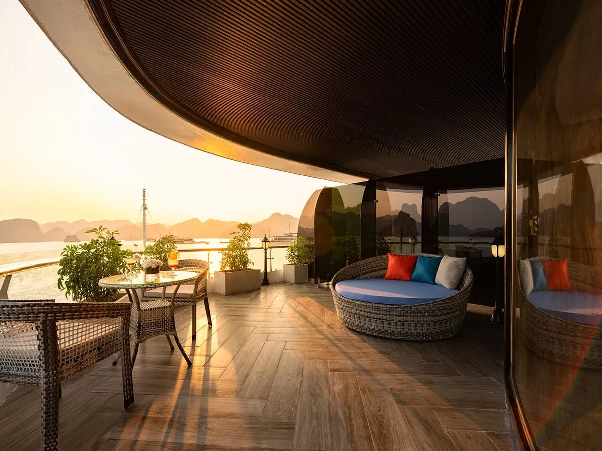 A curved private balcony with wicker chairs, a round table with drinks, a daybed with colourful cushions and potted plants, overlooking a bay with limestone karsts at sunset.