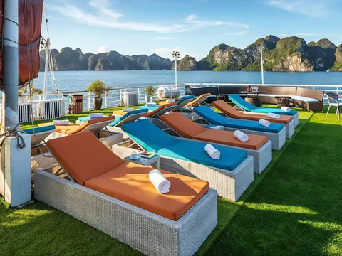 Rows of loungers on the turf sundeck beside a mast and an orange sail, with karst hills behind.