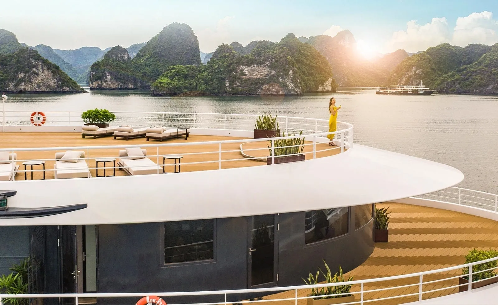 Curving upper sun decks of a modern dark-hulled cruise ship with white loungers, planters and a woman in a yellow dress, overlooking limestone karsts and another vessel in the bay.