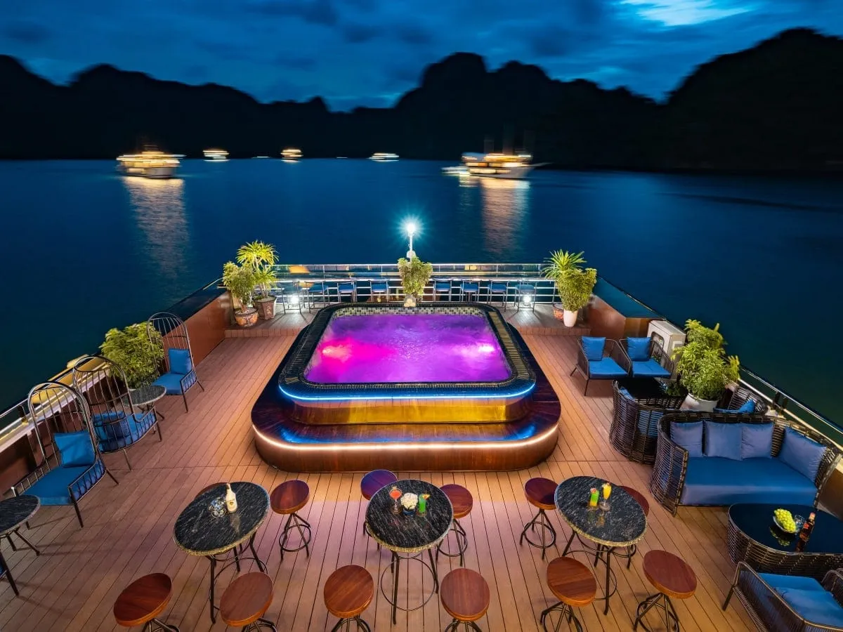 A dusk view from an upper deck of a cruise boat showing a lit purple jacuzzi surrounded by wooden decking, bar tables, stools, blue-cushioned seating and potted plants, with other lit boats anchored on the water and dark limestone karst silhouettes behind.