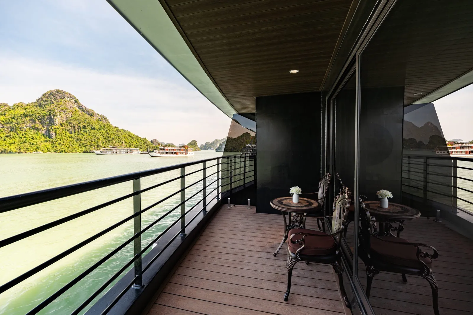 A private wooden-floored balcony with two small round tables, white flowers and cushioned metal chairs, overlooking green water, a limestone karst island and distant cruise ships.