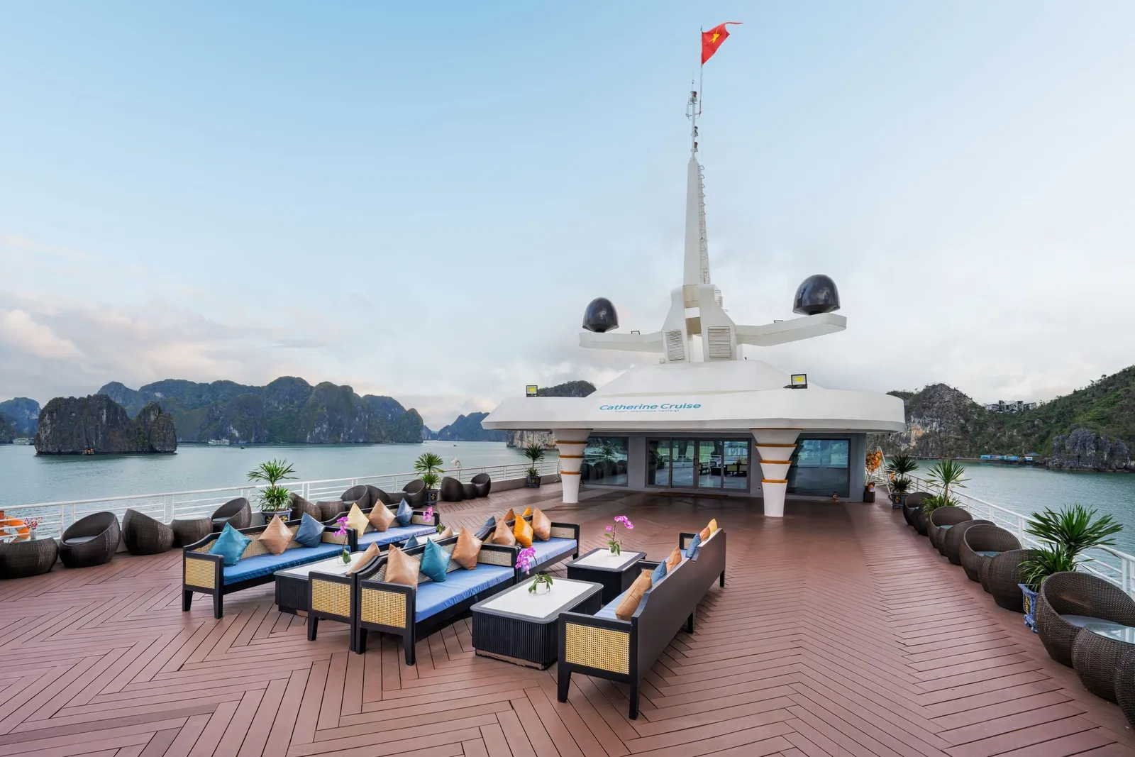 Sun deck of a cruise ship with blue-cushioned lounge sofas, wicker chairs and a wheelhouse structure flying a Vietnamese flag, with limestone karsts and calm water behind.