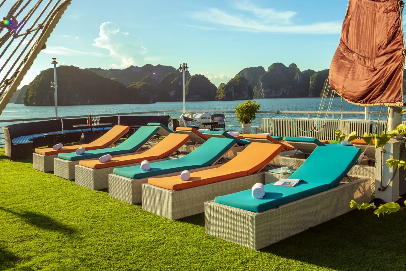 Loungers on the turf deck under a rigging line and an orange sail against a blue sky.