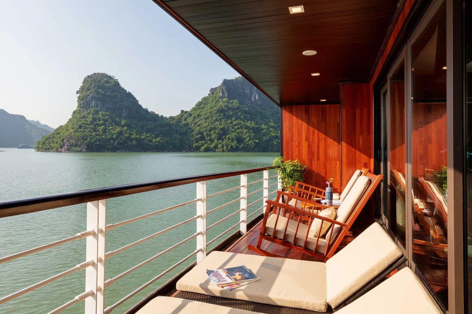 A private wooden balcony with rocking chairs and cushioned loungers beside a white railing, overlooking green water and forested limestone karst islands.