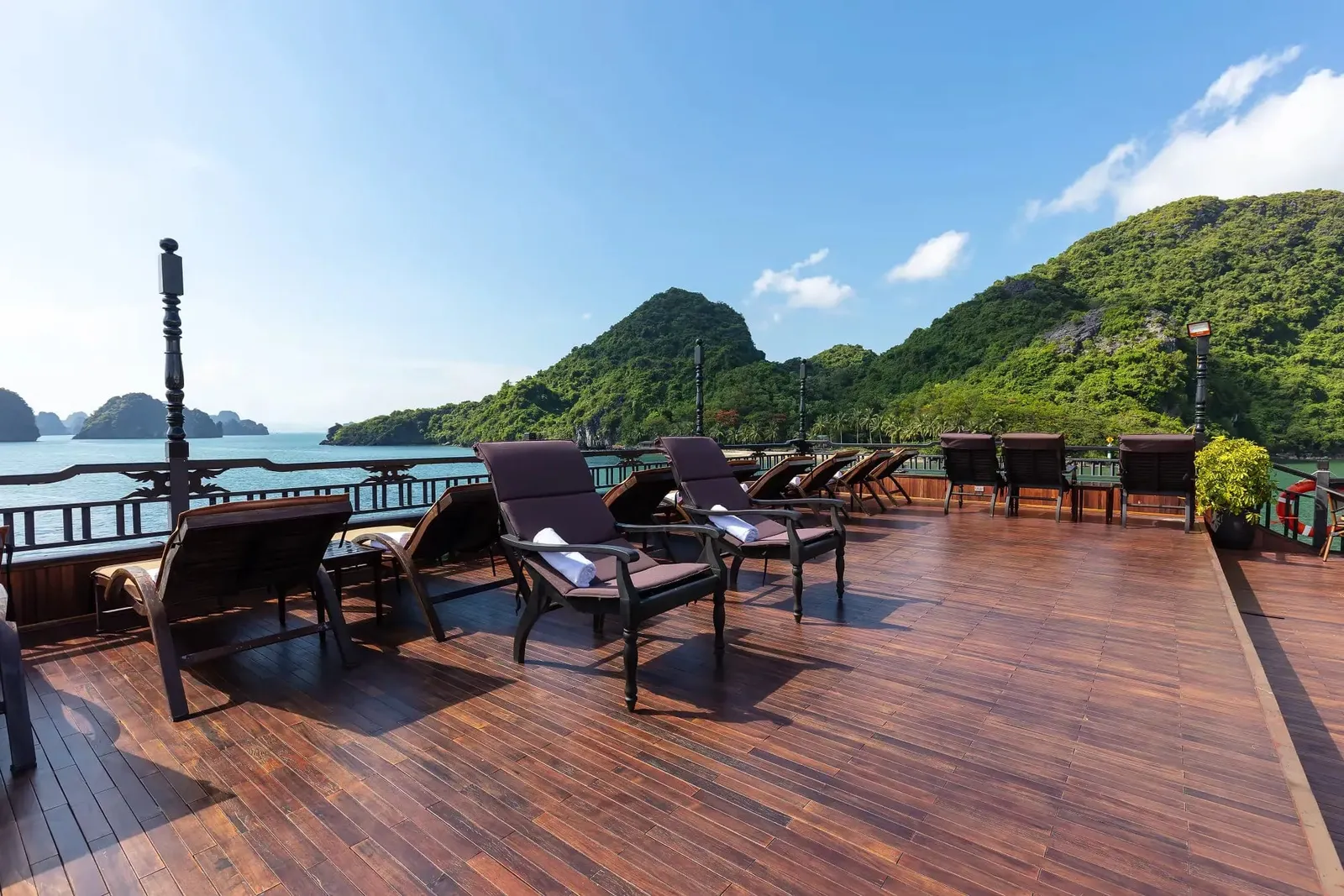 A wooden sundeck with dark loungers and chairs with towels, a railing, and green limestone karst islands across calm water under a blue sky.