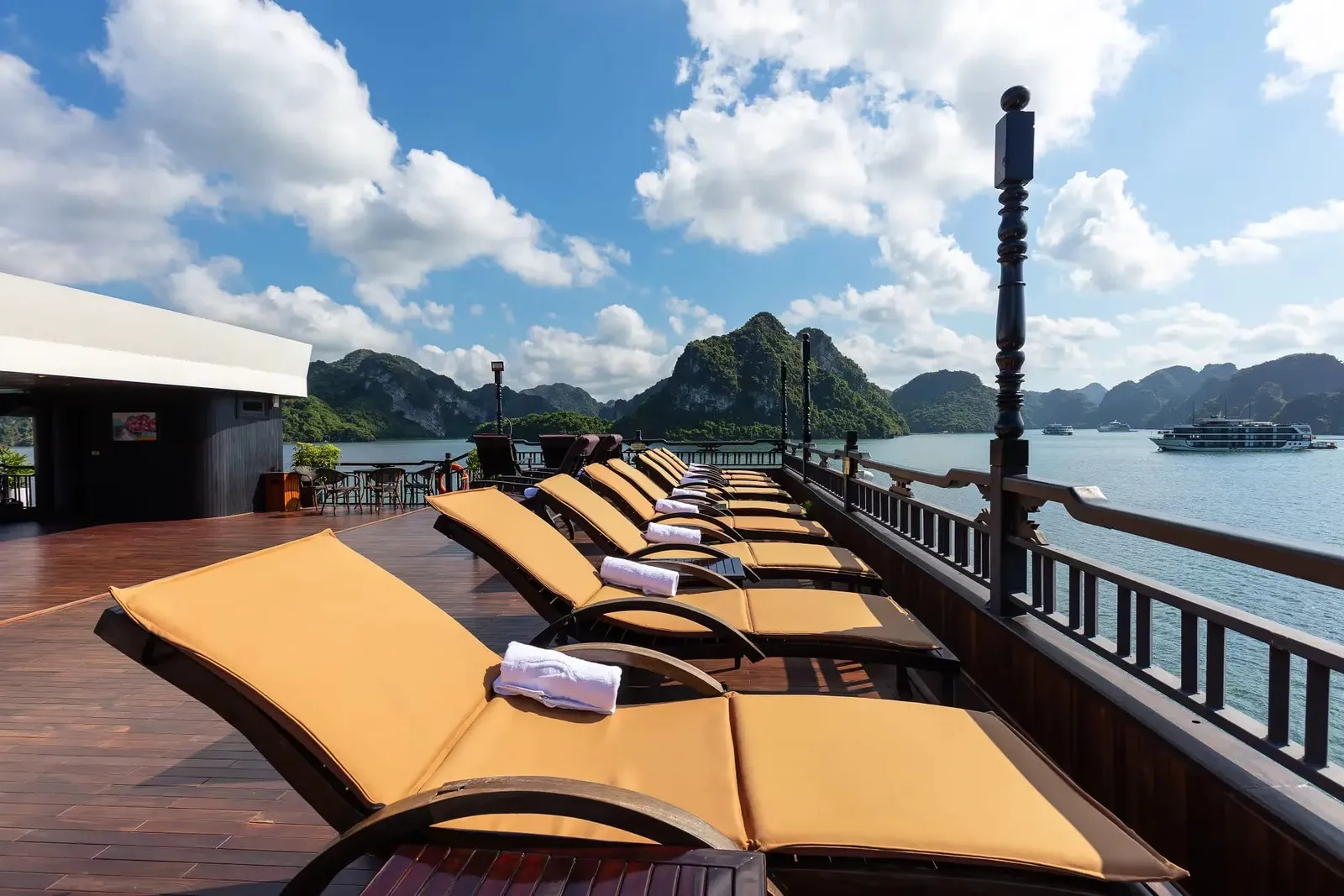 A wooden sun deck with a row of orange-cushioned loungers topped with folded white towels, a dark wooden railing, and limestone karst islands with another cruise ship in the distance under a blue sky.