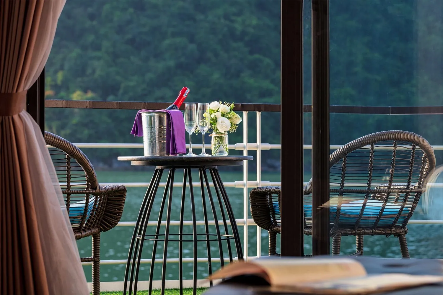 A private balcony seen through glass doors, with two wicker chairs, a small bistro table holding a champagne bottle in an ice bucket, two glasses and white flowers, with green water and forested hills beyond the railing.