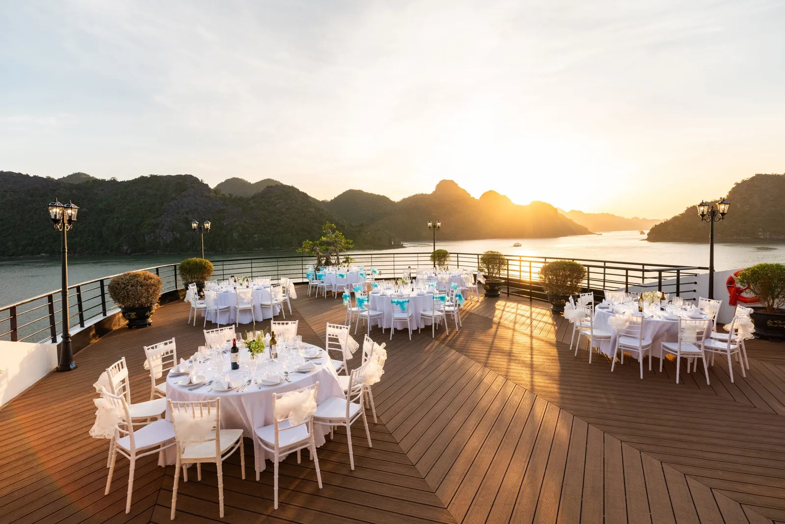 A wooden sun deck set with round white-clothed dining tables and white chairs, with lamp posts and potted shrubs, overlooking karst islands in calm water at sunset.