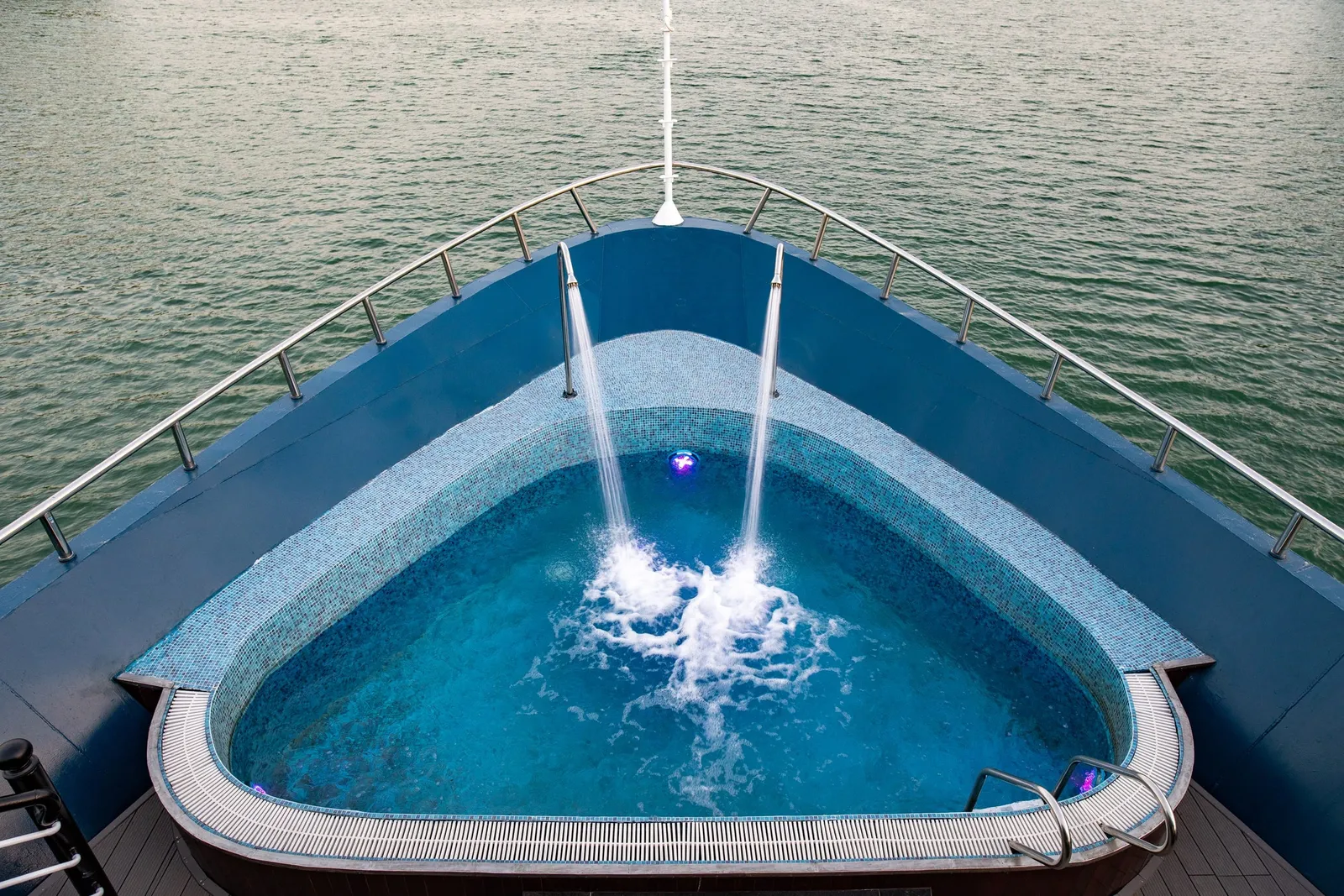 A triangular jacuzzi pool with water jets on the bow deck of a cruise boat surrounded by calm greenish water.