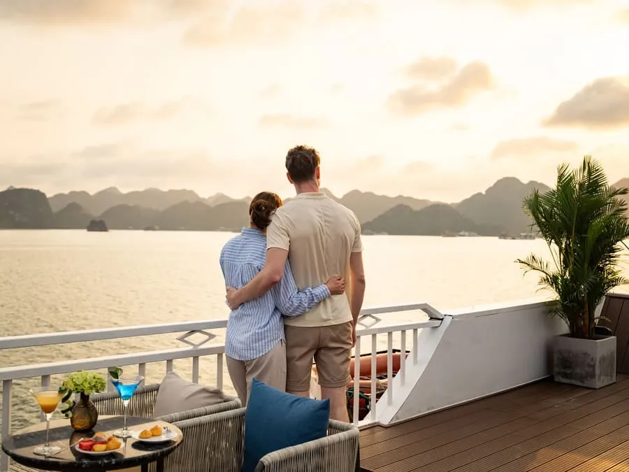 A couple seen from behind at the deck railing with cocktails and fruit, looking out over the bay at sunset.