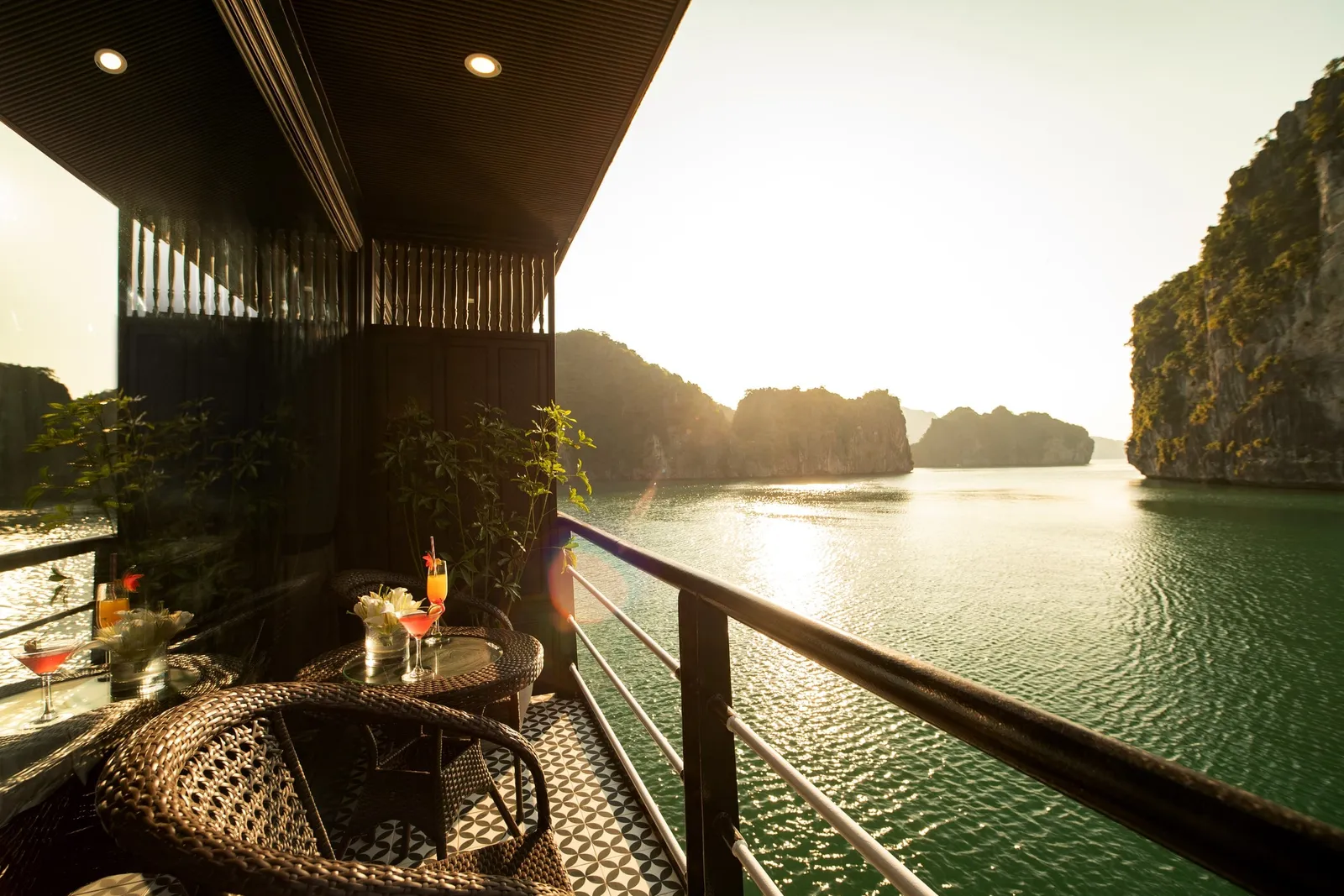 A balcony deck with wicker chairs, a small table with cocktails and flowers, and a railing overlooking green water and limestone karst islands at sunset.