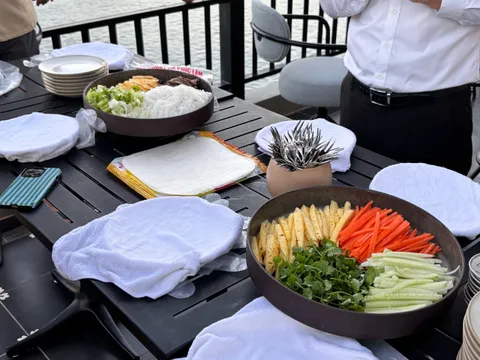 A deck table set up for a spring-roll making session with bowls of sliced pineapple, carrot, cucumber, herbs, noodles, rice paper and folded towels, two staff standing by a railing with calm water and a white cruise boat behind.