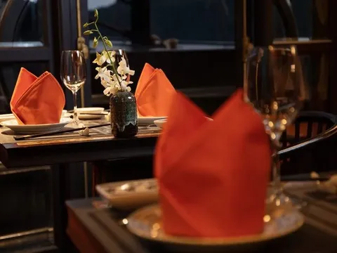 Dark wooden dining tables set with folded orange napkins, wine glasses, plates, chopsticks and a small vase of white orchids, in a dimly lit room with window frames behind.