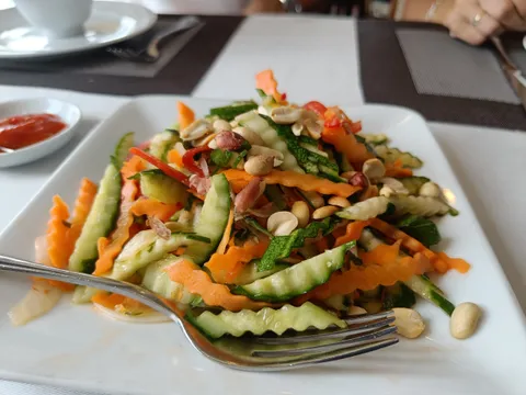 A plated vegetable salad with peanuts served on a dining table with place settings and a cup in the background.
