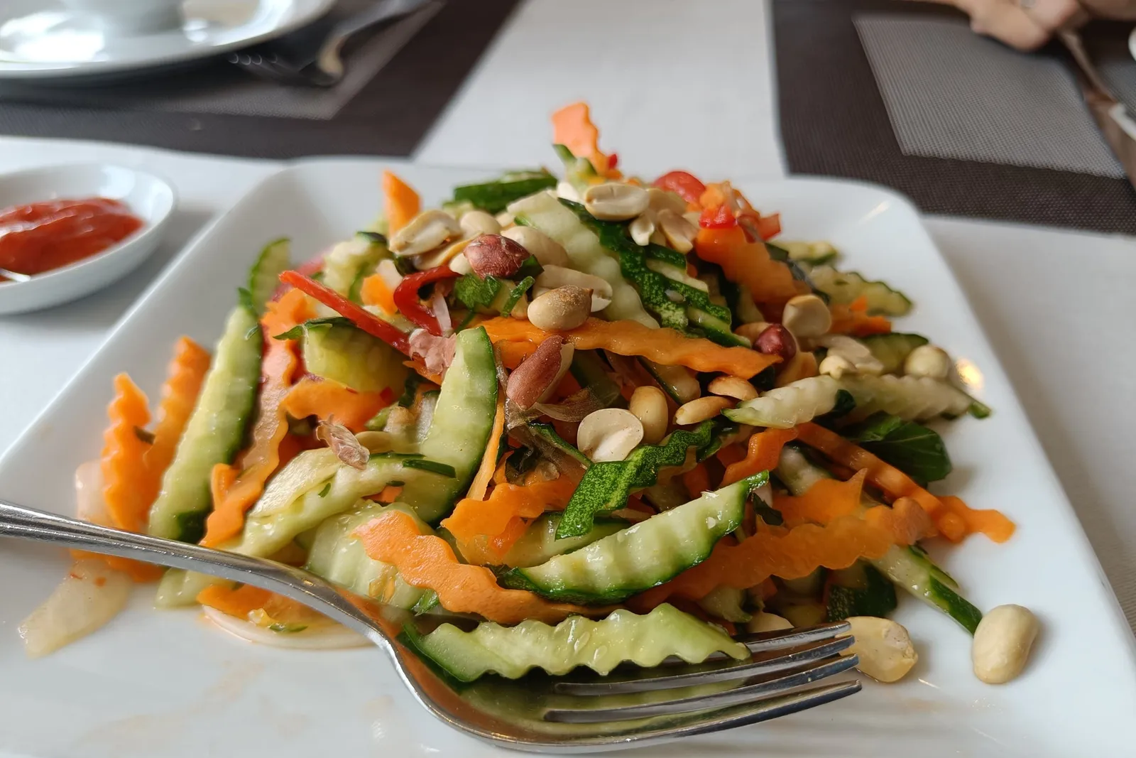 A plated vegetable salad with peanuts served on a dining table with place settings and a cup in the background.
