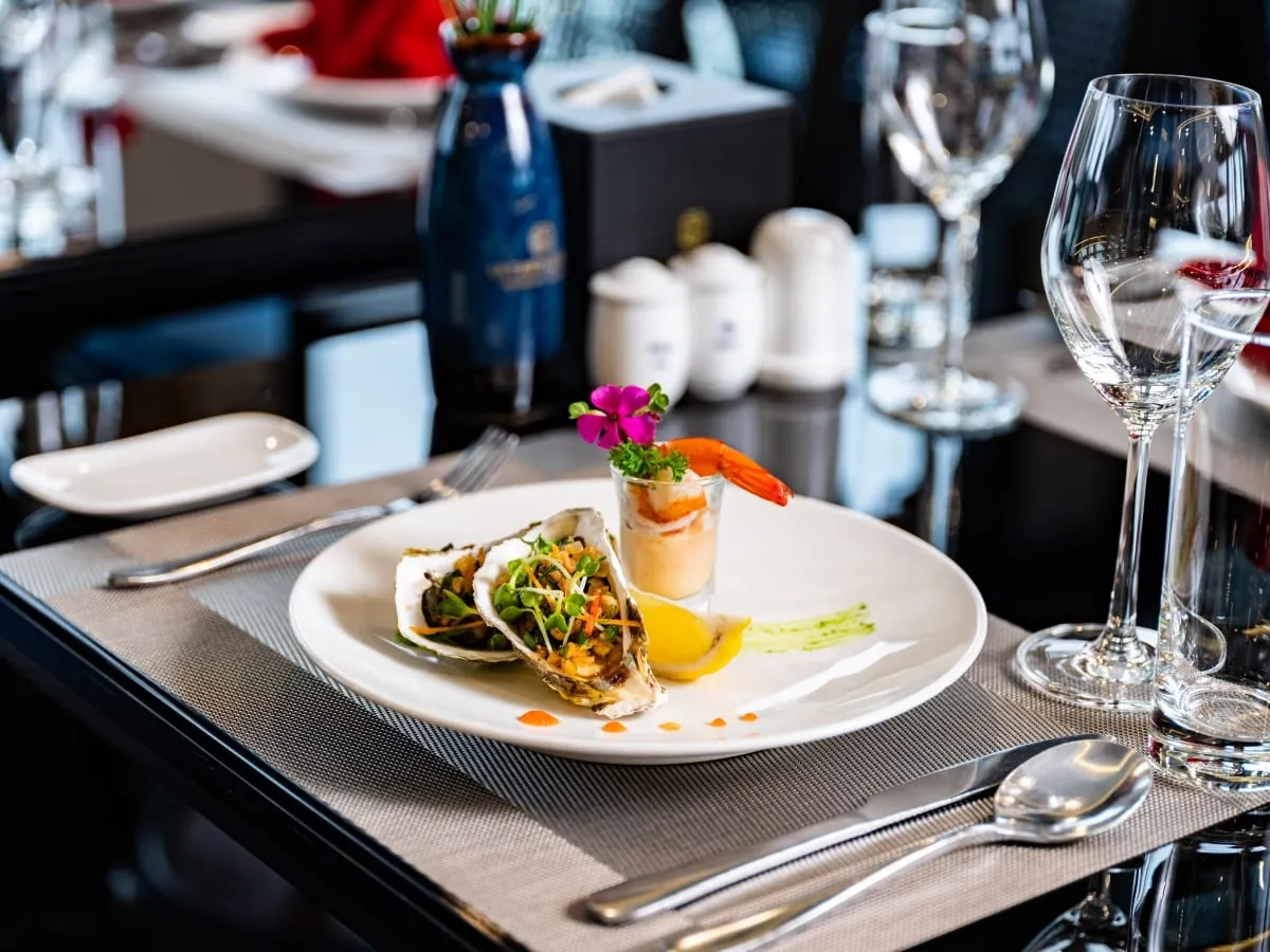 A plated appetizer of two baked oysters with greens and a shrimp cocktail shot glass sits on a place setting with wine glasses, cutlery and a blue branded ceramic bottle on a dark table.