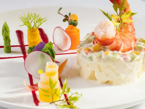 An artfully plated appetizer with shrimp, vegetable garnishes, and decorative carved vegetables on a white square plate.