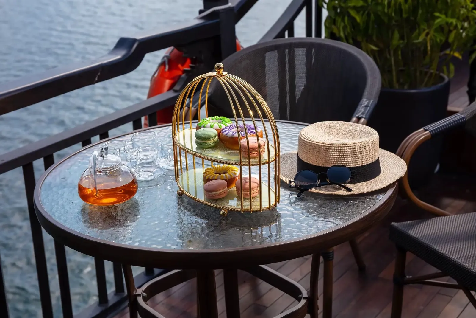 A round glass-top table on a wooden balcony deck with a gold birdcage-style tray of macarons and donuts, a glass teapot with tea, glasses, a straw hat with sunglasses, and water visible beyond the railing.