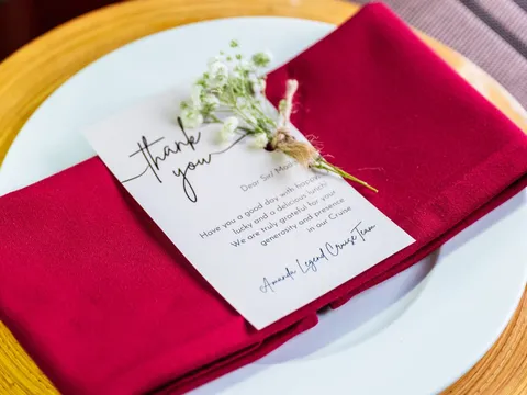 A folded red napkin on a white plate and yellow woven charger, topped with a printed thank-you card signed 'Amanda Legend Cruise Team' and a sprig of baby's breath.