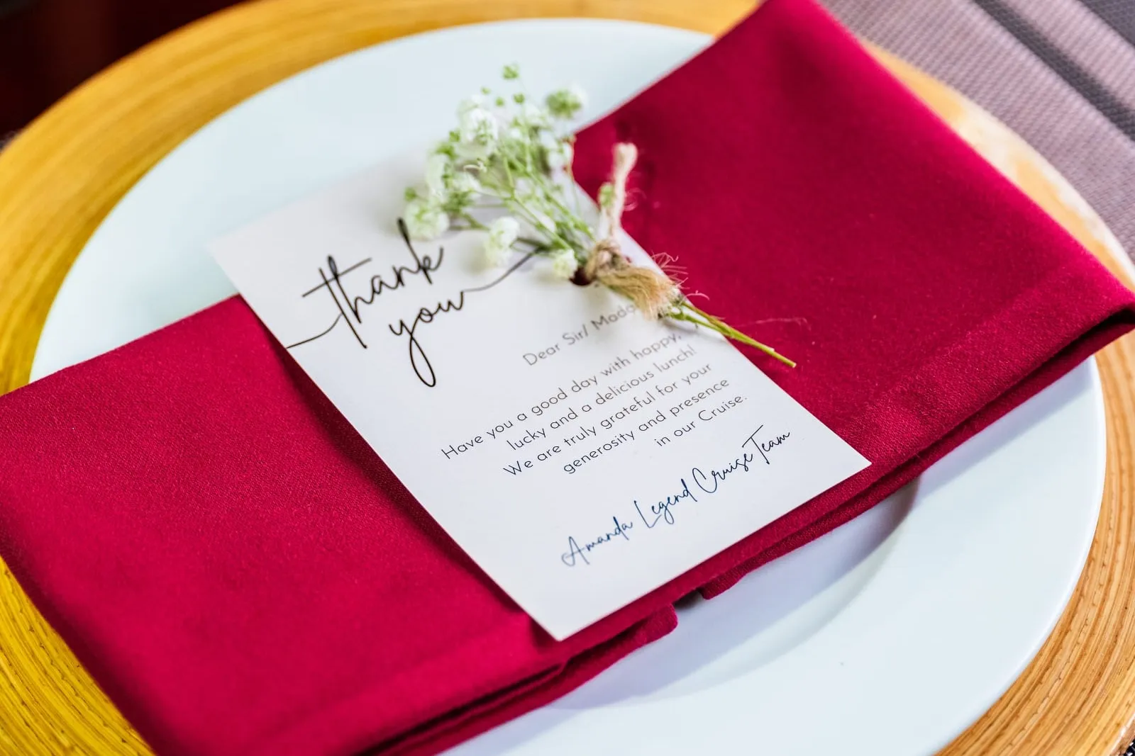 A folded red napkin on a white plate and yellow woven charger, topped with a printed thank-you card signed 'Amanda Legend Cruise Team' and a sprig of baby's breath.